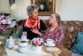 Eine Malteser Helferin sitzt gemeinsam mit einer älteren Dame auf einem Sofa und die beiden unterhalten sich angeregt.
