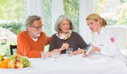 Auf dem Foto sind drei Personen zu sehen, die hinter einem Tisch sitzen. Eine Frau in Malteserkleidung zeigt den beiden anderen Personen, d.h. einem älteren Pärchen, das Hausnotrufgerät. 