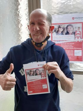 Ein regelmäßiger Gast des Kölner Wohlfühlmorgens hält das aktuelle Plakat in den Händen. Er ist froh über das neue Angebot „Warm durch den Winter“. Foto: Malteser Köln