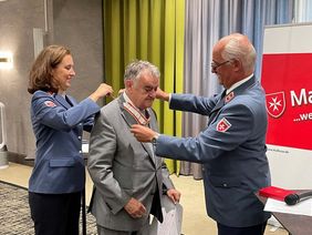 Die Regional- und Landesgeschäftsführerin der Malteser, Dr. Sophie von Preysing, und der Regional- und Landesbeauftragte, Rudolph Herzog von Croÿ, legen Minister Herbert Reul das Kommandeurkreuz an.